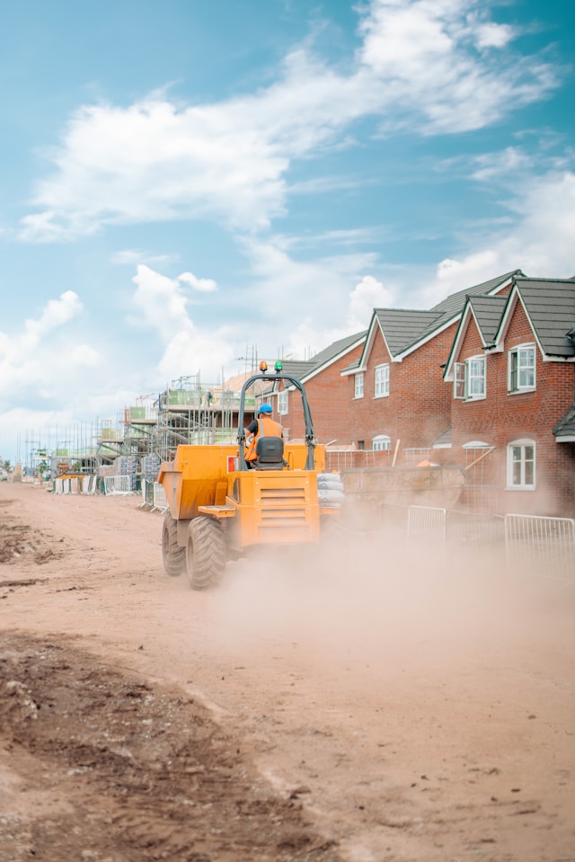 A building contractor drives yellow heavy machinery down a temporary road next to a construction site of new build houses.